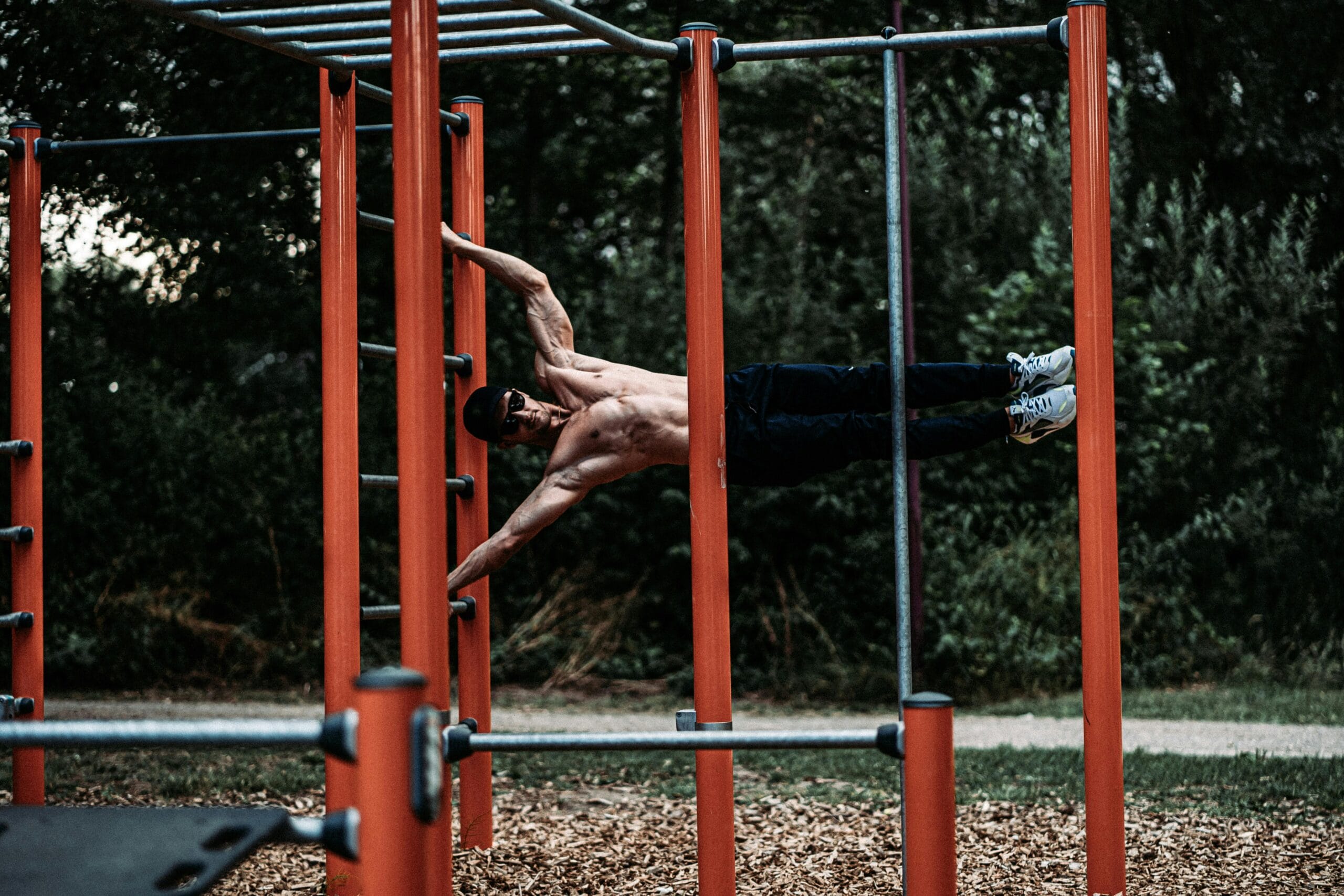 A shirtless man demonstrates strength and agility on outdoor calisthenics bars.